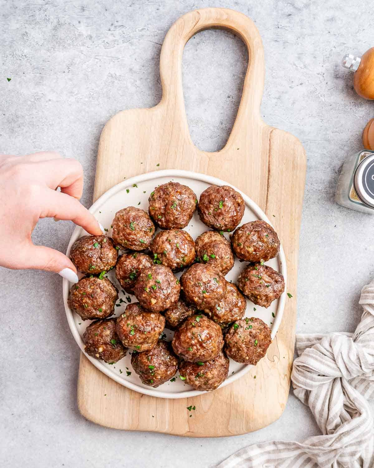 A hand reaching for a meatball on a white round plate filled with meatballs on a cutting board.